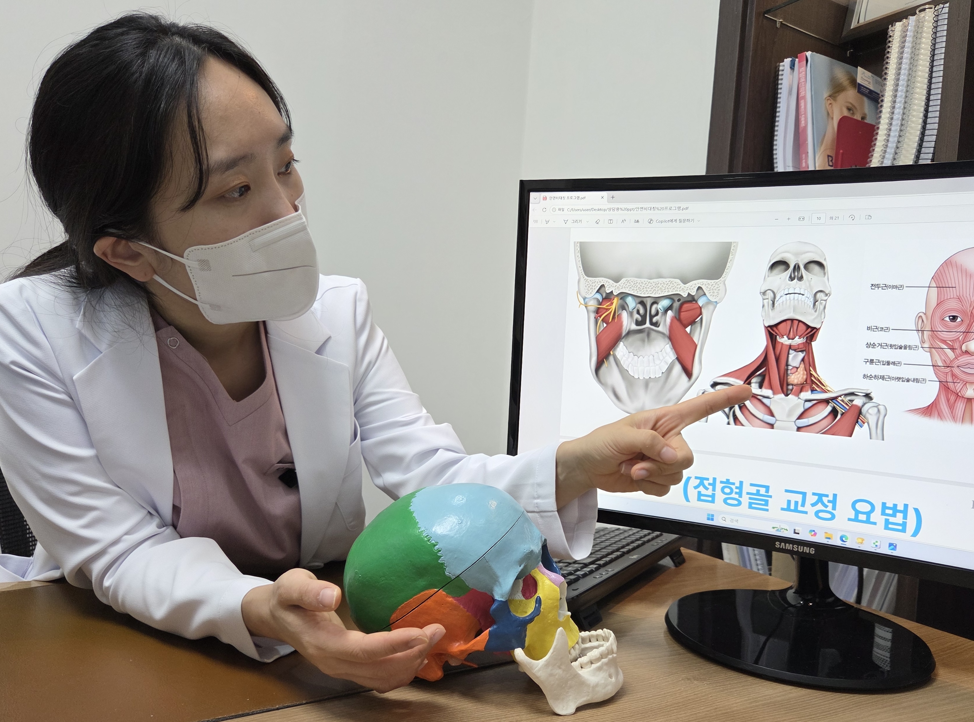 원장이 두개골 모형을 들고 모니터의 안면 근육 자료를 가리키며 설명하는 모습